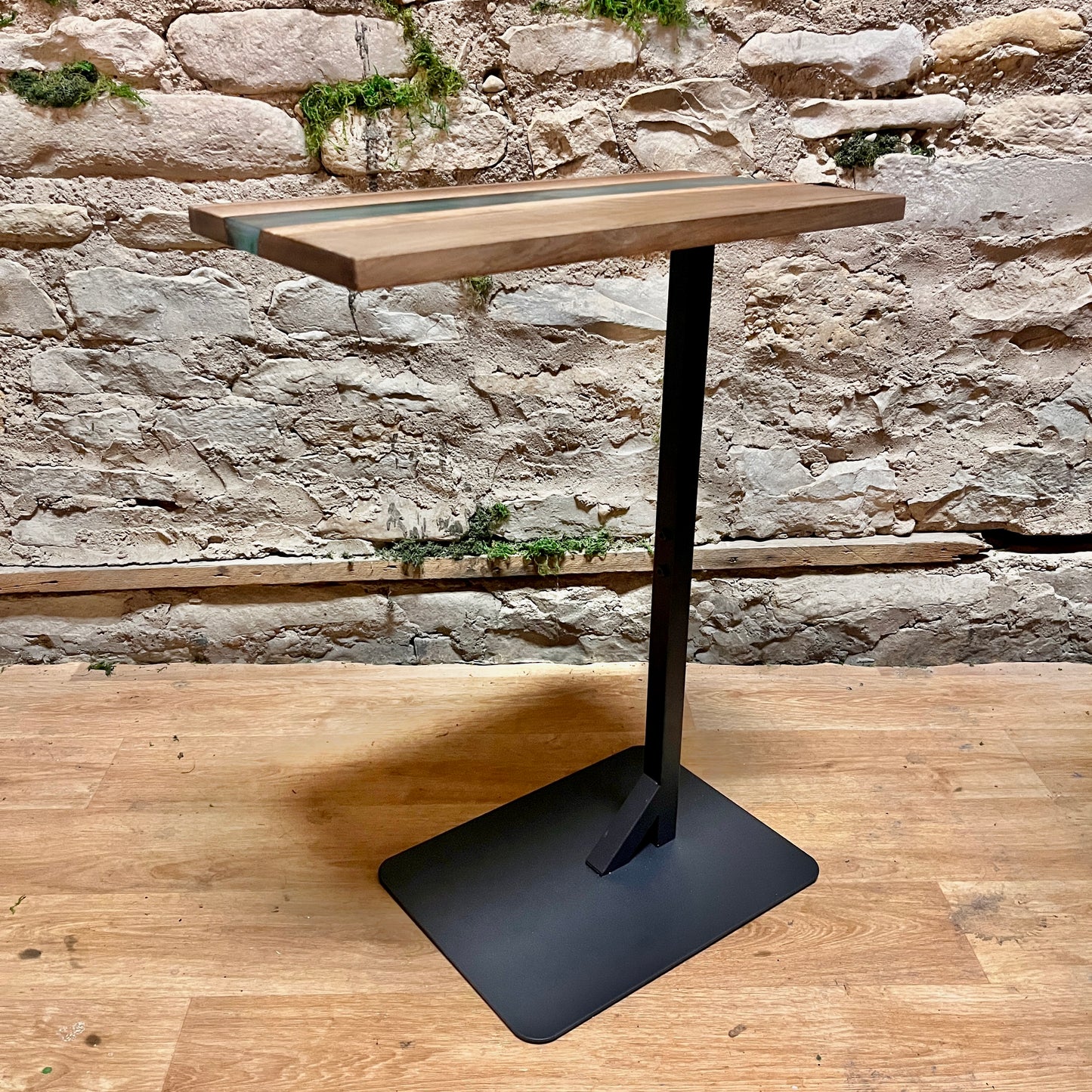 Wooden epoxy river side table with live edge wood and a black metal base against a stone wall.