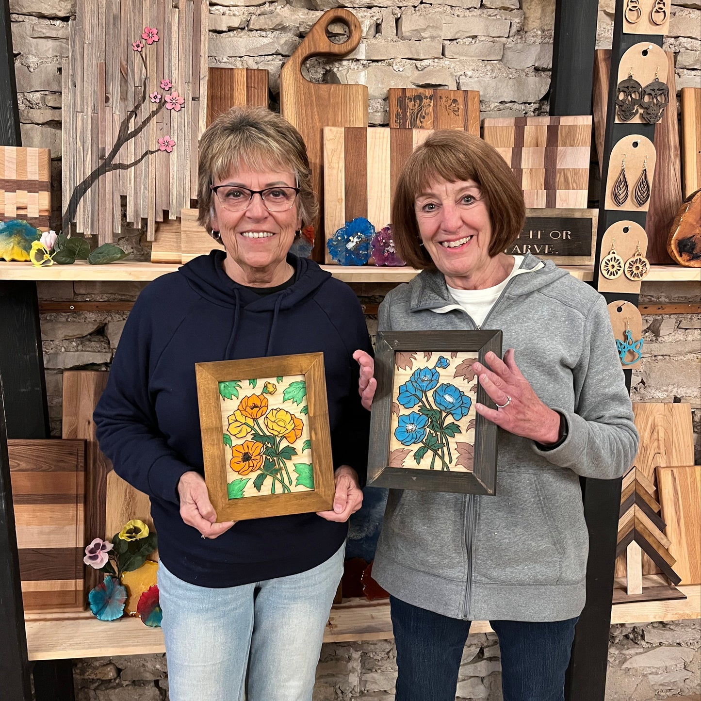 Two adults showcasing their framed artwork creations at a workshop setting, with one depicting orange flowers and the other showing blue flowers.