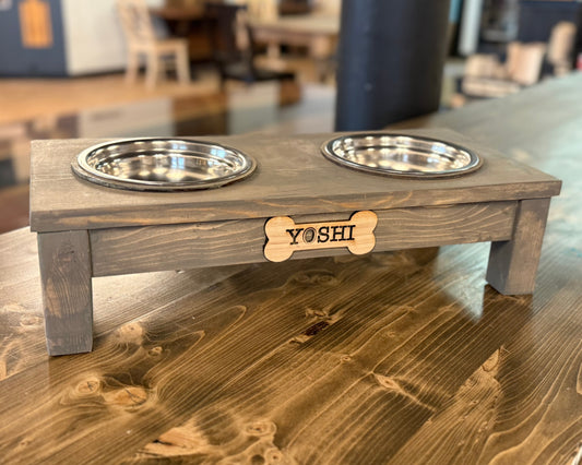 Wooden pet feeding station with two dog bowls and a bone-shaped wood piece withe the name 'Yoshi' on a wooden table.