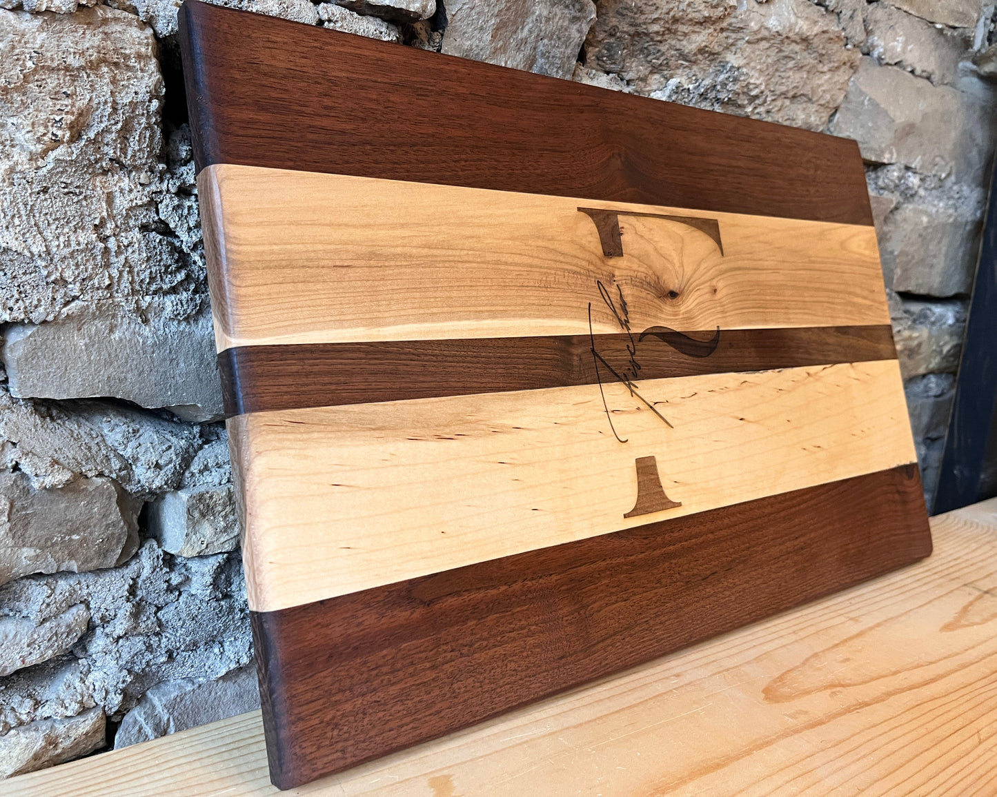 Wooden cutting board with an engraved last name and initial and unique grain pattern against a stone wall background