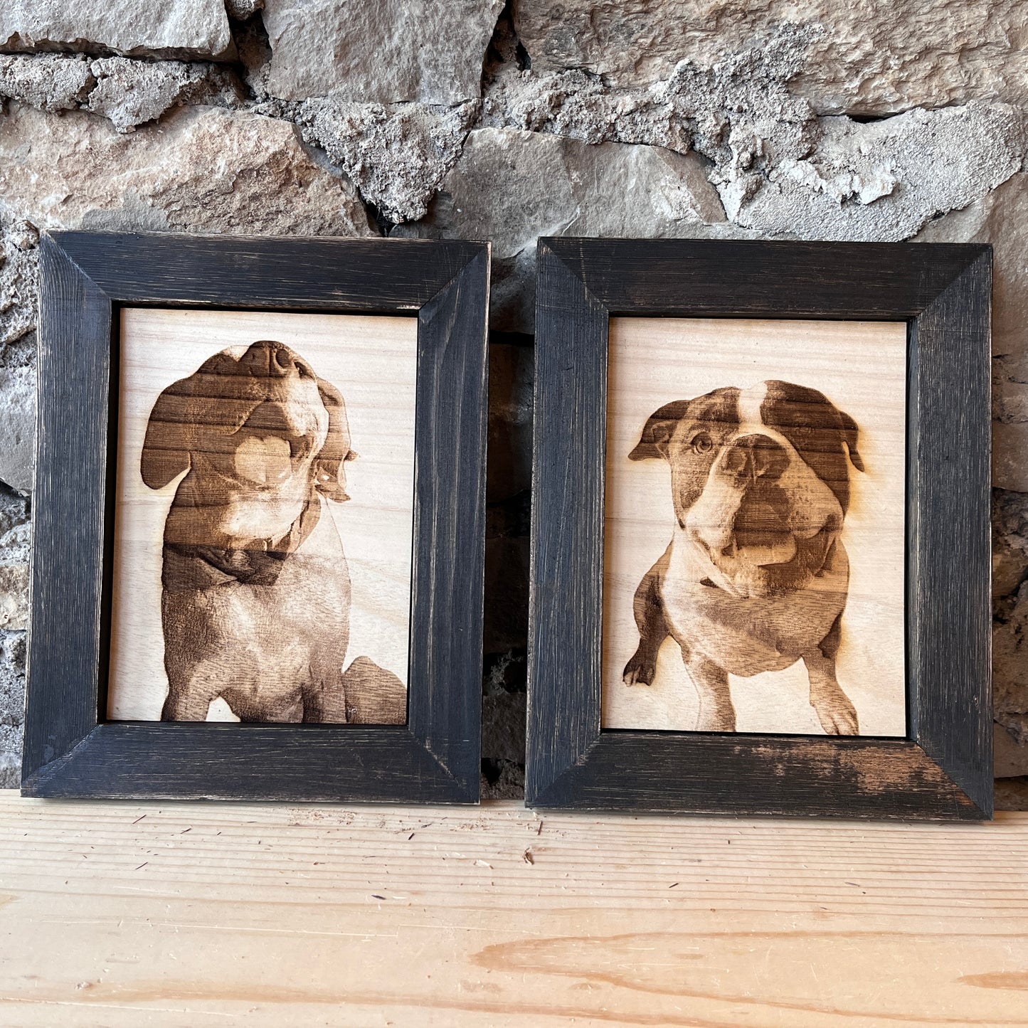 Two framed laser engraved portraits of dogs on wood, displayed against a stone wall on a wooden surface.