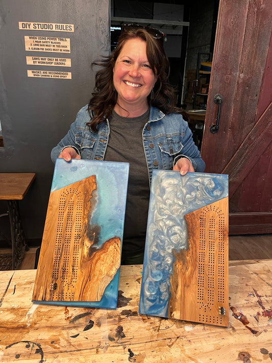 Person holding two wooden epoxy river cribbage board crafts with drilled holes in a workshop setting.