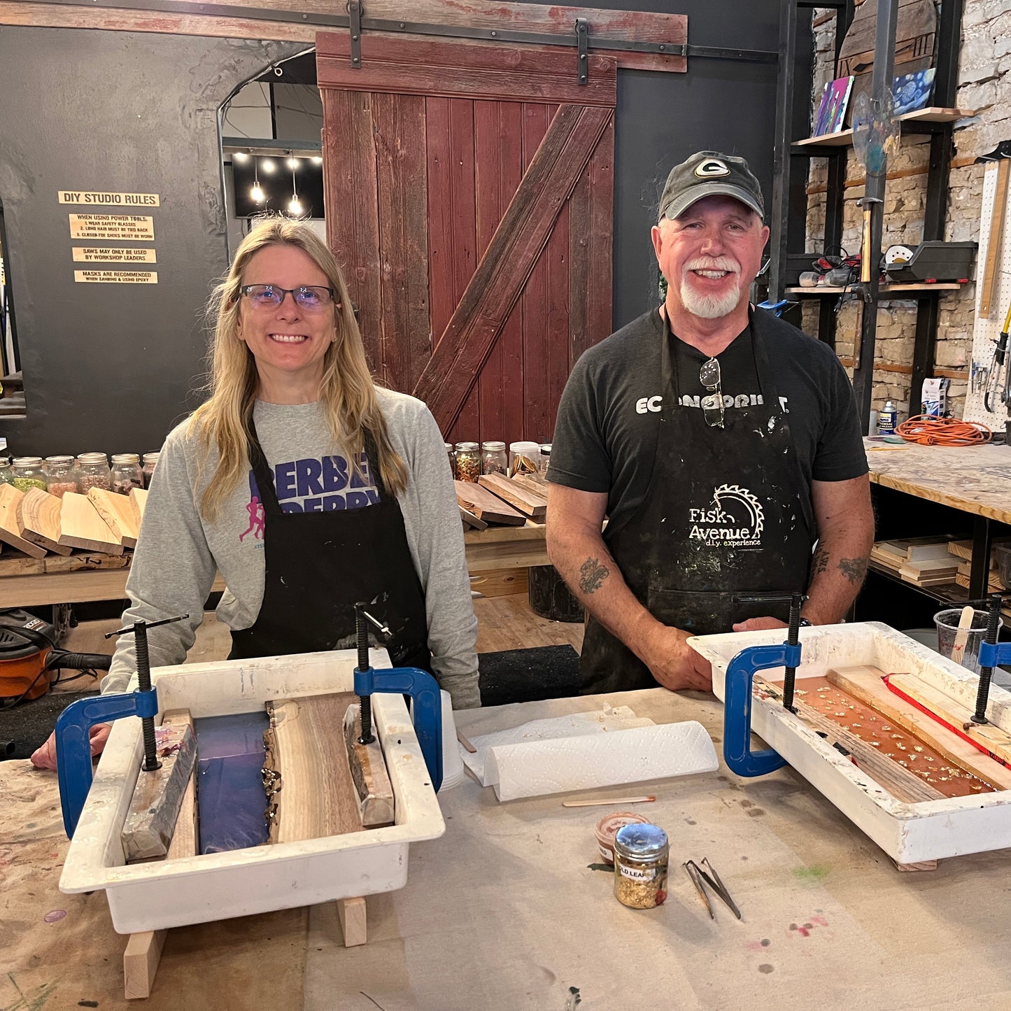 Two people in a workshop setting making epoxy river serving trays with live edge wood in molds on workbenches.