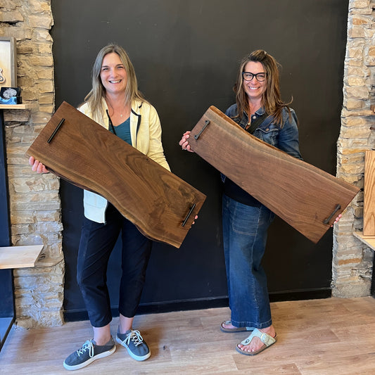 Two people holding large wooden live edge charcuterie boards in a studio with stone walls and wooden flooring.