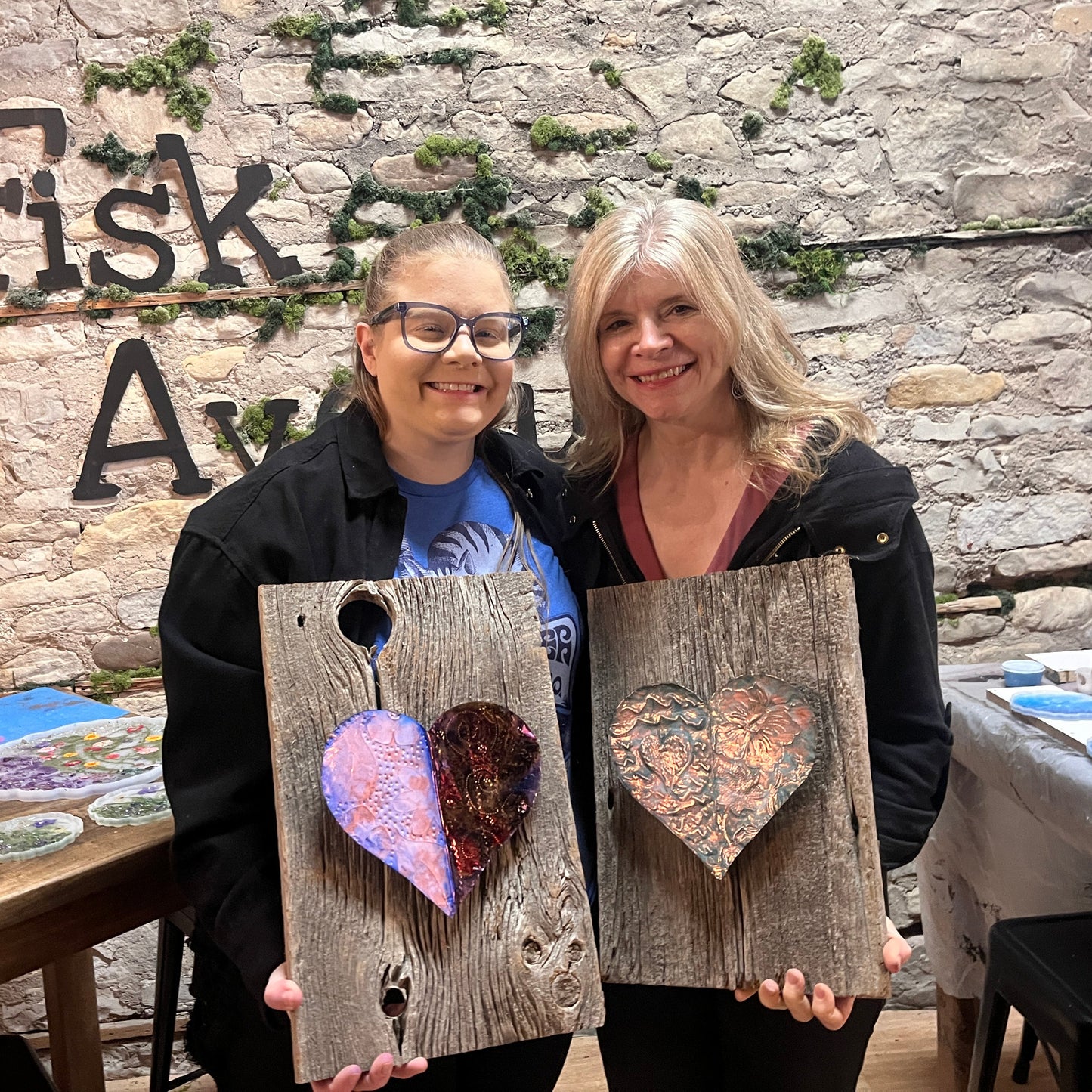 Two women holding rustic wooden boards with embossed copper heart designs in front of a stone wall with 'Fisk Avenue' text.