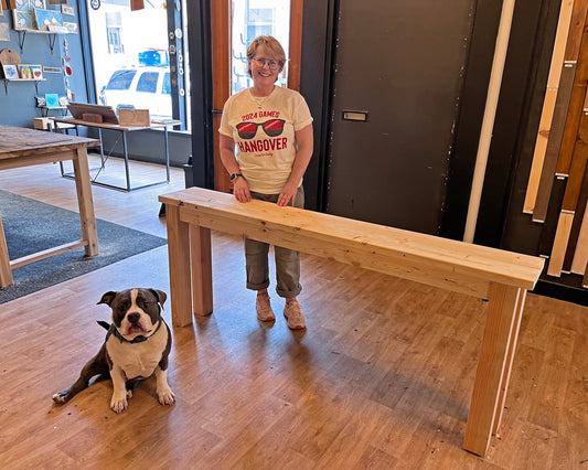 Woman standing next to a wooden sofa table with a dog sitting on the floor in a studio setting.