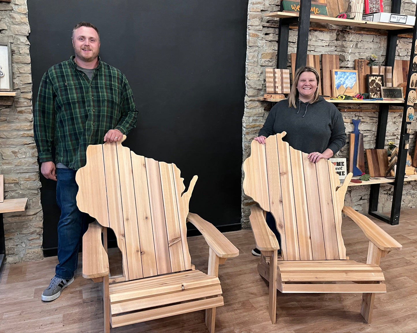 Two people standing with two wooden Adirondack outdoor chairs shaped like the state of Wisconsin in a store setting.