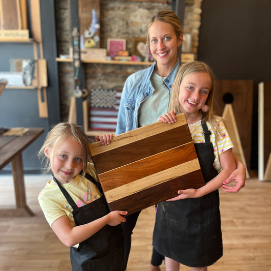 Two young girls and a woman holding a wooden cutting board in a workshop setting.