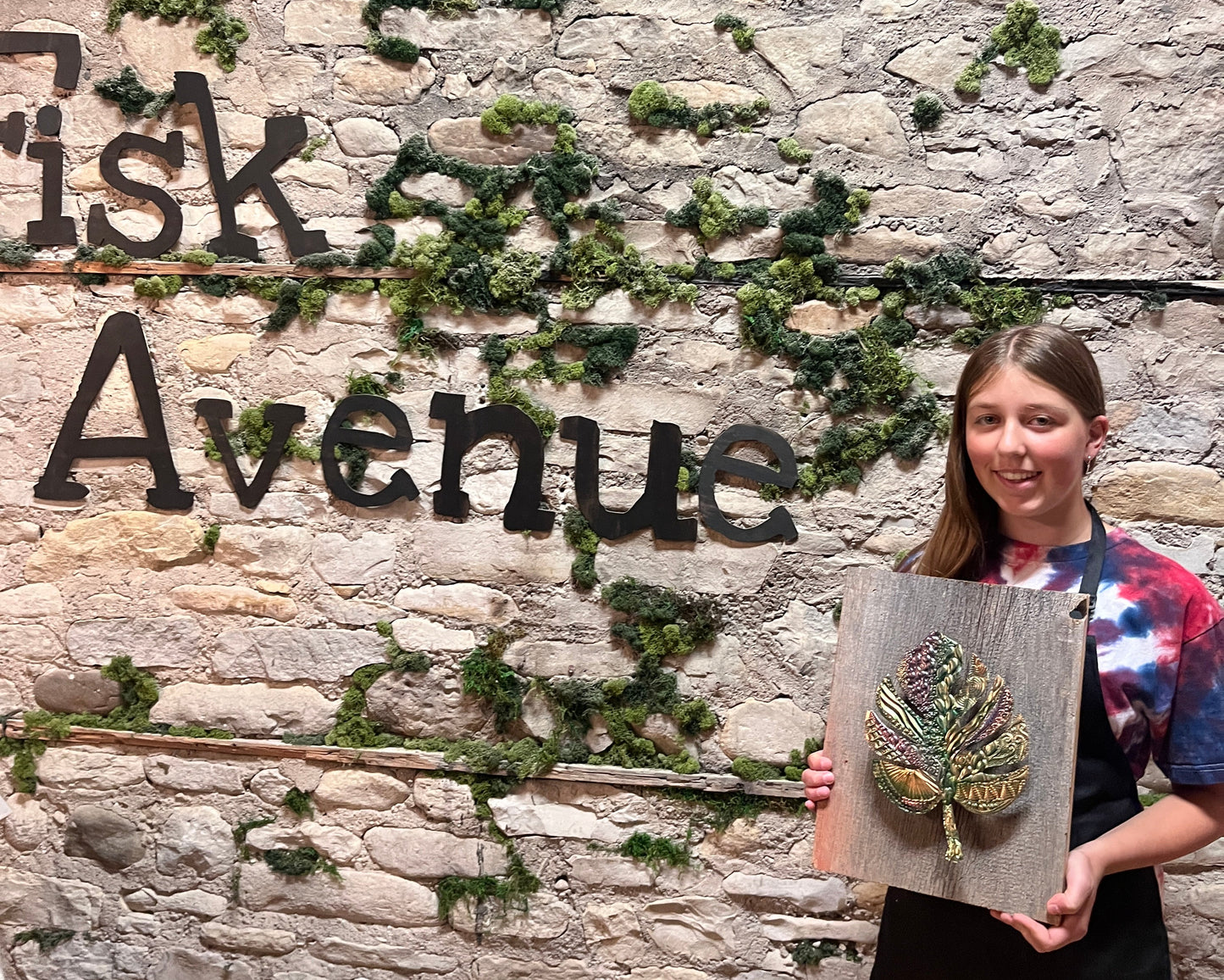 Person holding a framed copper leaf artwork against a stone wall with 'Fisk Avenue' text.