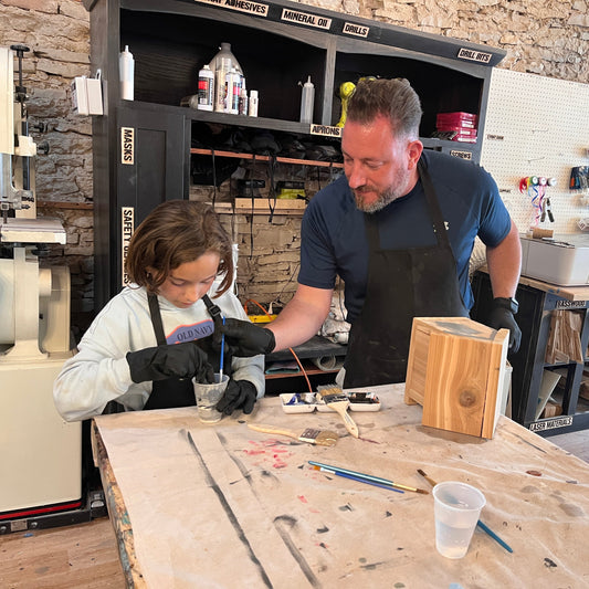 Man and child working on a birdhouse craft painting project in a workshop.