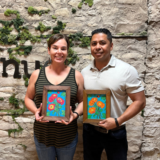 Two people holding framed paintings of flowers against a stone wall.