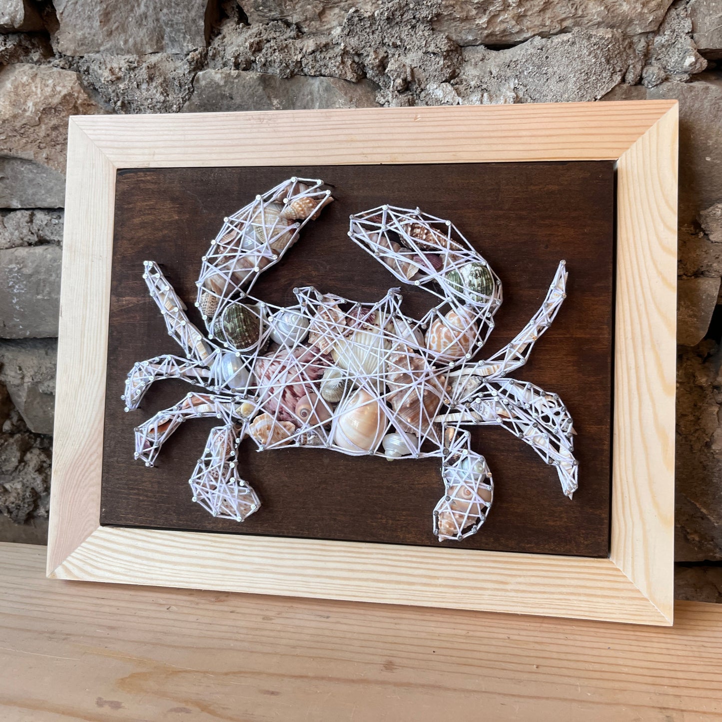 Framed artwork of a crab made from string, wood, and shells against a stone wall.
