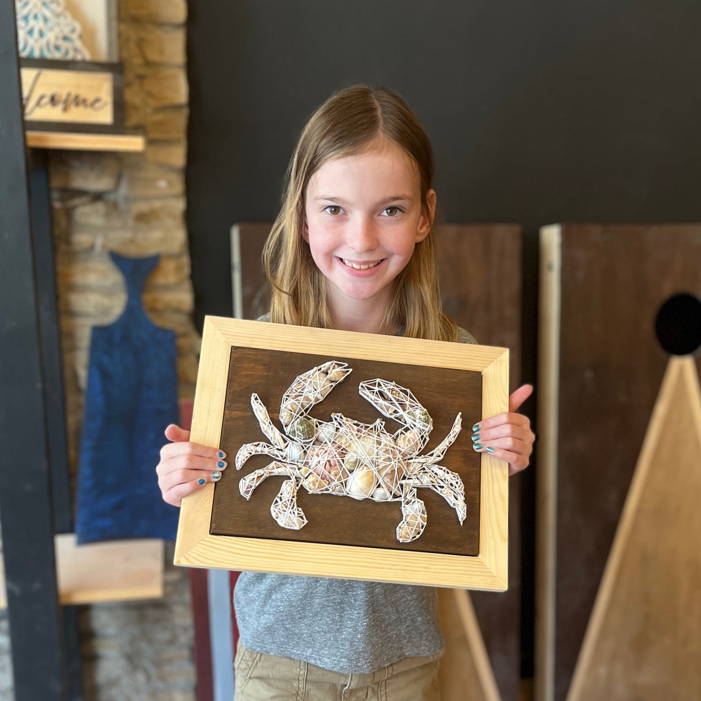 Young girl holding a framed string art piece of a a crab with shells in a studio setting.