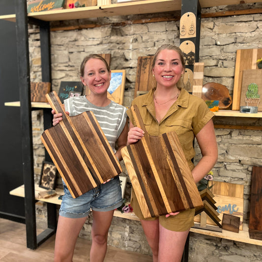 Two women holding wooden pizza peel boards in a studio setting with shelves and decor in the background.