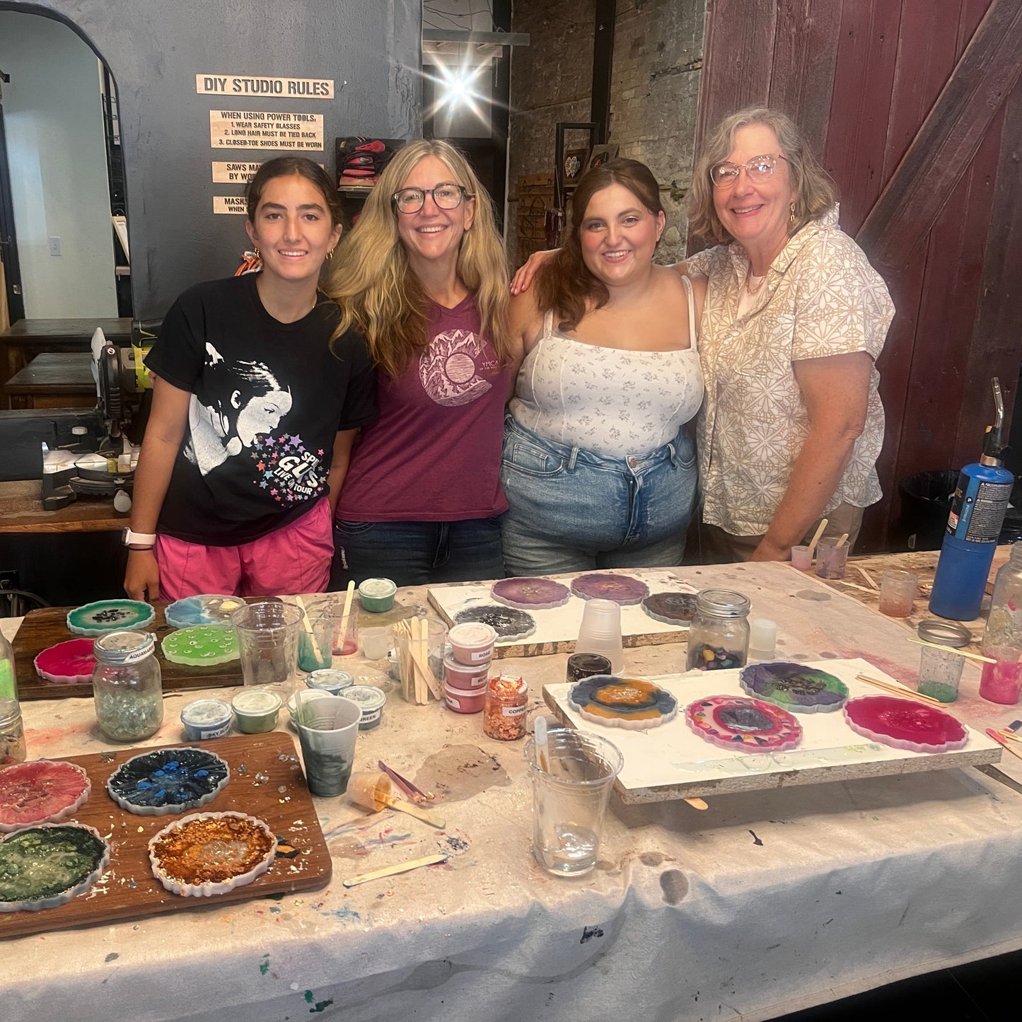 Four people standing in a studio with epoxy art supplies and coasters on a table.