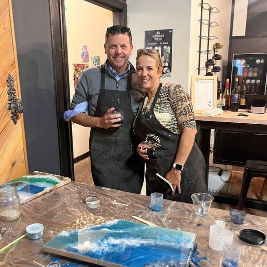 Two people in aprons standing behind a table with epoxy tray supplies, smiling at the camera.