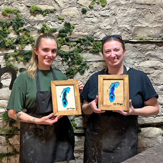 Two women holding framed Lake Michigan artworks in front of a stone wall with 'Fisk Avenue' sign.