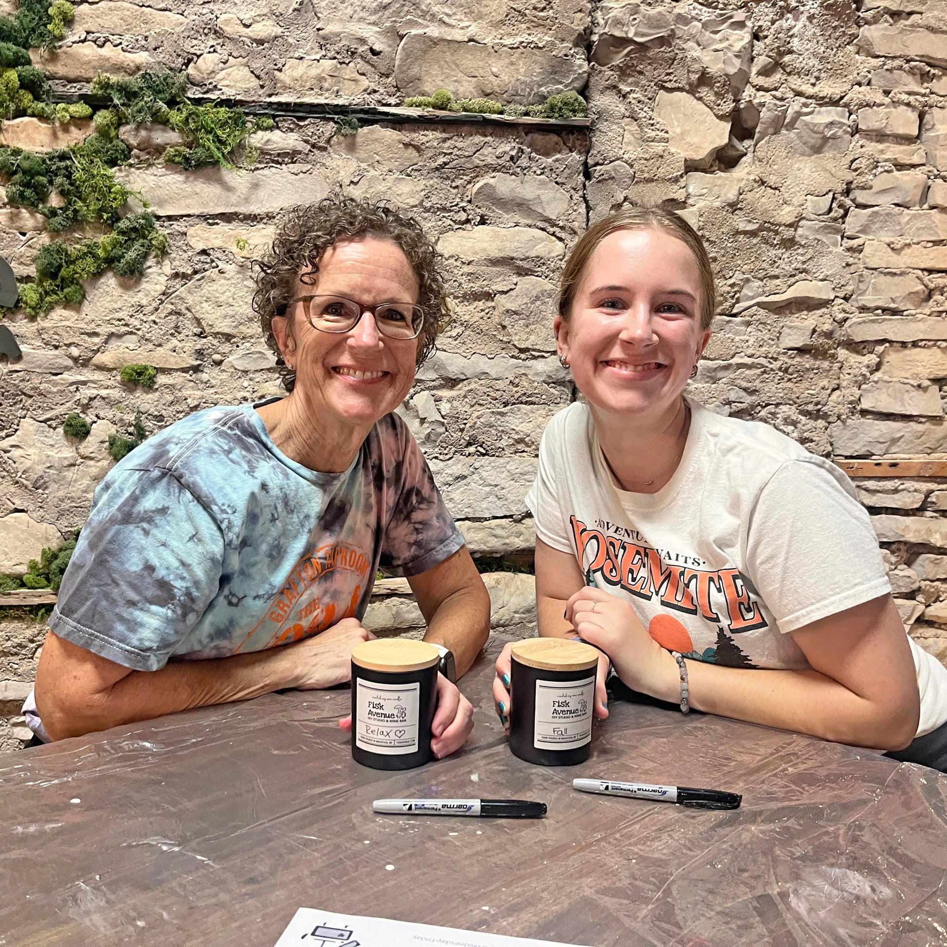 Two people sitting at a table with candles and markers against a stone wall.