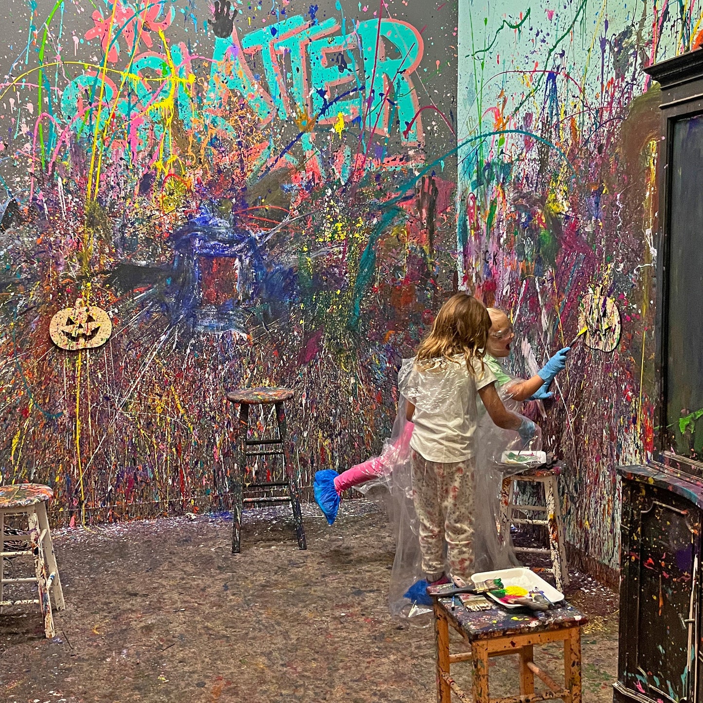Splatter room with colorful graffiti on walls and a person painting a wooden jack-o-lantern.