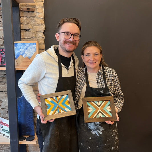 Two people holding framed wood mosaic artworks in a casual studio setting