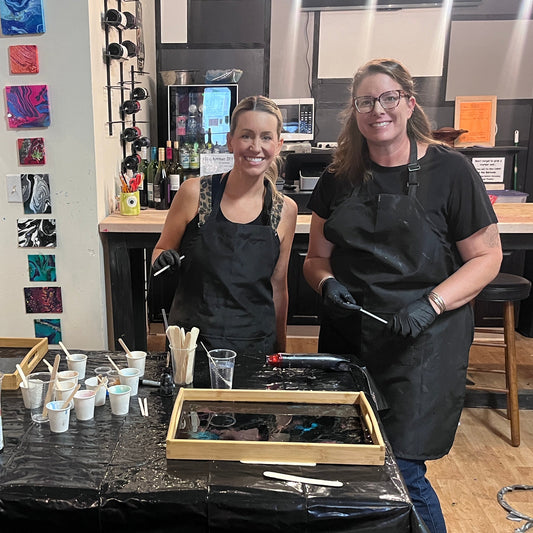 Two women wearing black aprons stand behind a table covered with small cups, epoxy, and wooden trays inside a studio with wood walls and shelves in the background.