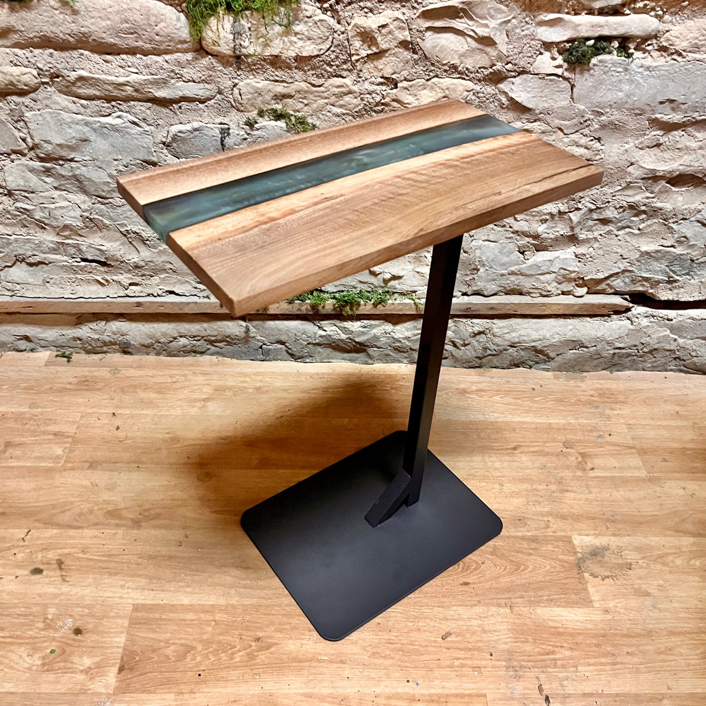 Wooden epoxy river side table with live edge wood and a black metal base against a stone wall.