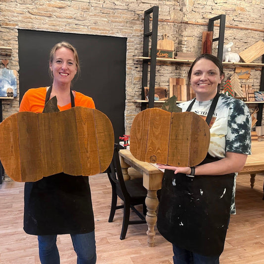 Two women holding wooden pumpkin decorations in a workshop setting.