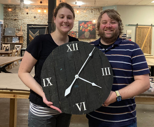 Two adults holding a round wooden clock with numerical Roman numerals and a wooden texture finish.