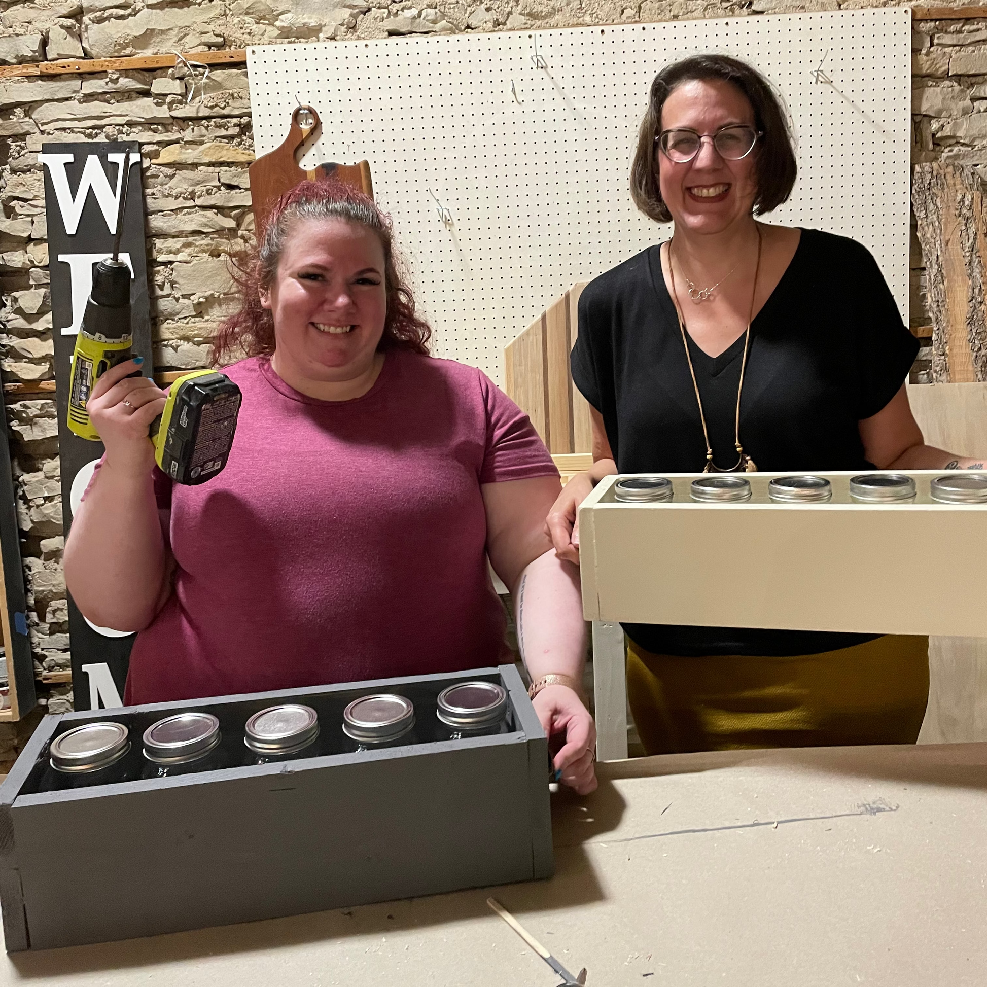Two women standing with a drill and centerpieces boxes by a stone wall background