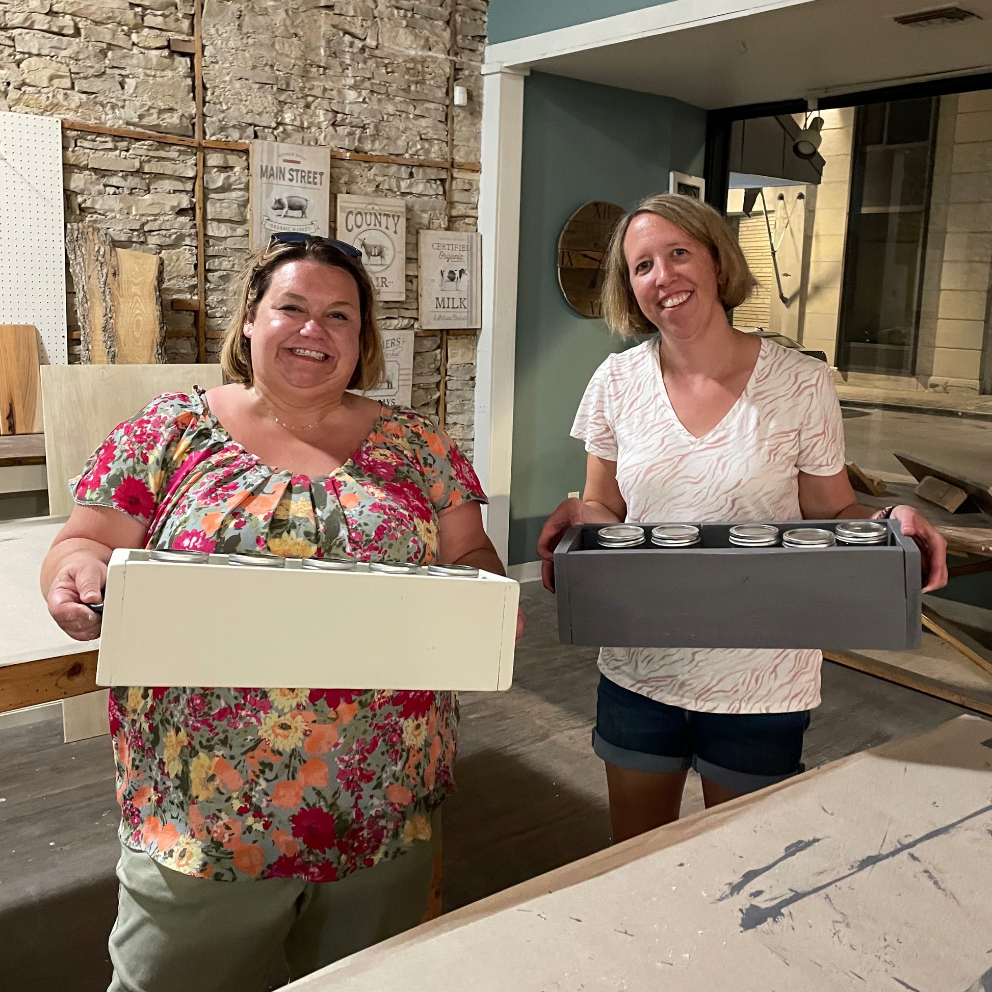 Two women holding centerpiece boxes in a room with stone walls and a blue wall.