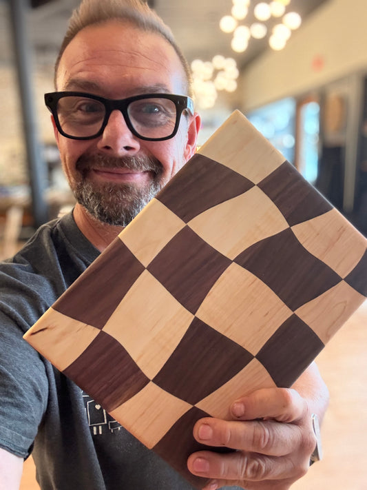 A man with glasses and a beard holds up a square wooden board with a dark and light checkerboard pattern in a shop setting.