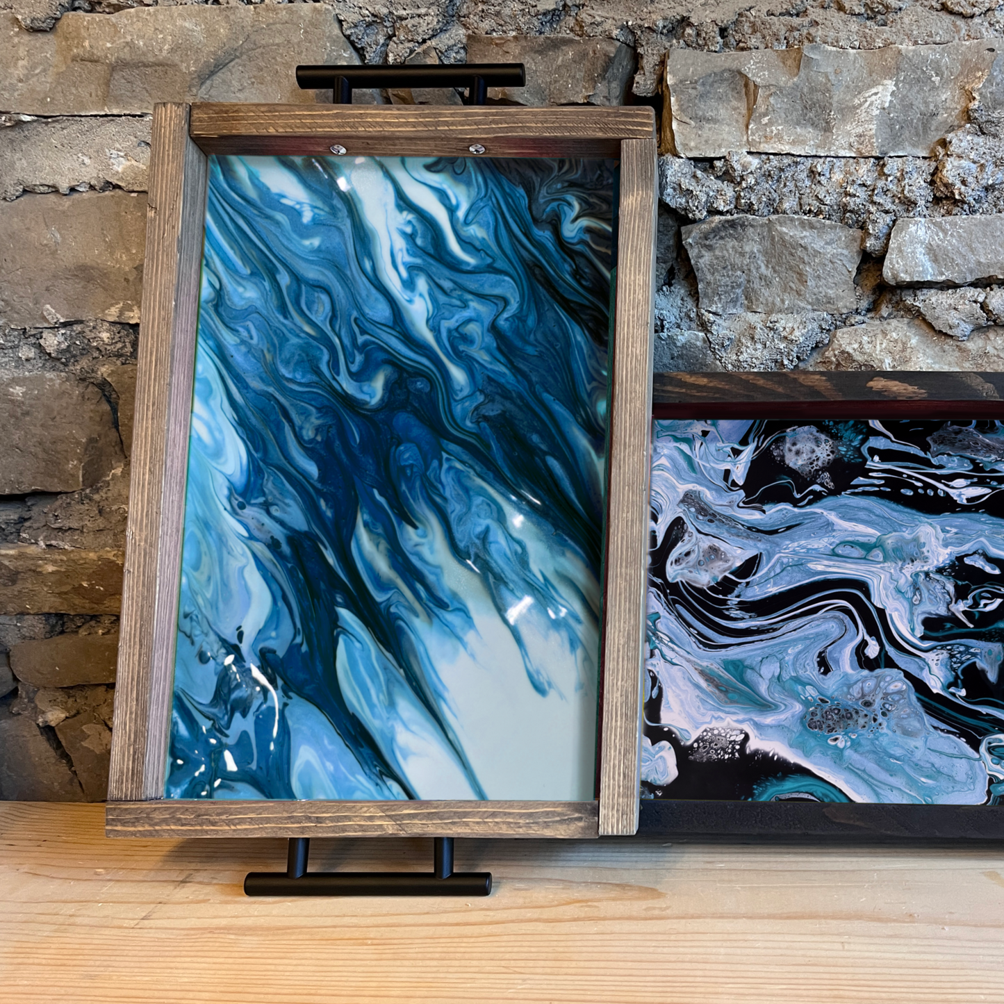 Two acrylic pour trays with abstract designs with blue swirls on a wooden surface with a stone wall background.