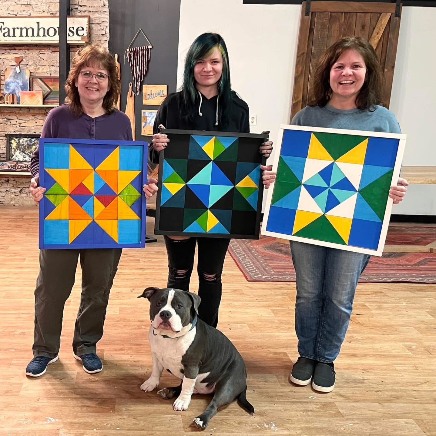 Four women holding colorful wooden quilt art with a dog in the foreground in a room with wooden floors and stone walls.