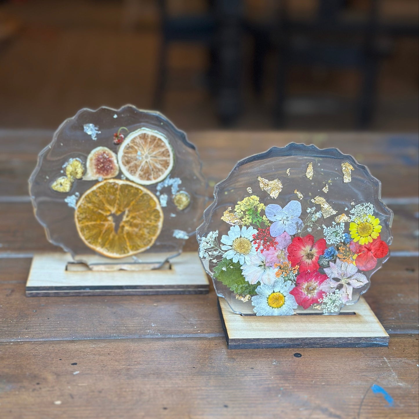 Two epoxy geode tabletop decor pieces with pressed flowers and oranges with wooden stands on a wooden surface.