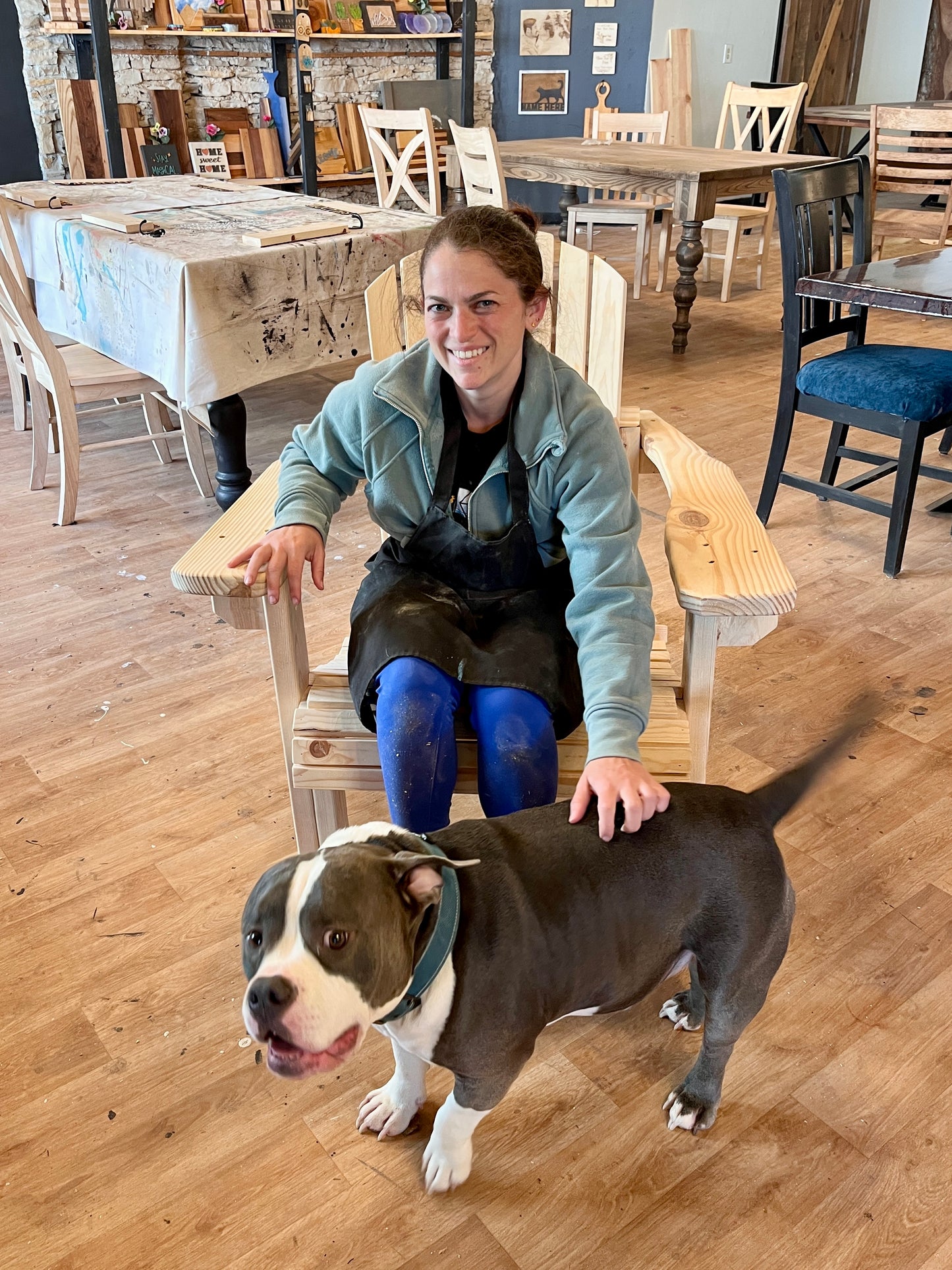 Woman in apron sitting in a wooden Adirondack chair in a workshop with a dog.