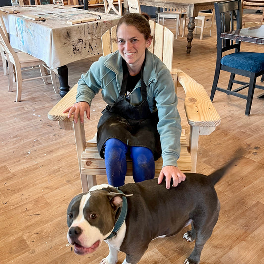 Woman in apron sitting in a wooden Adirondack chair in a workshop with a dog.