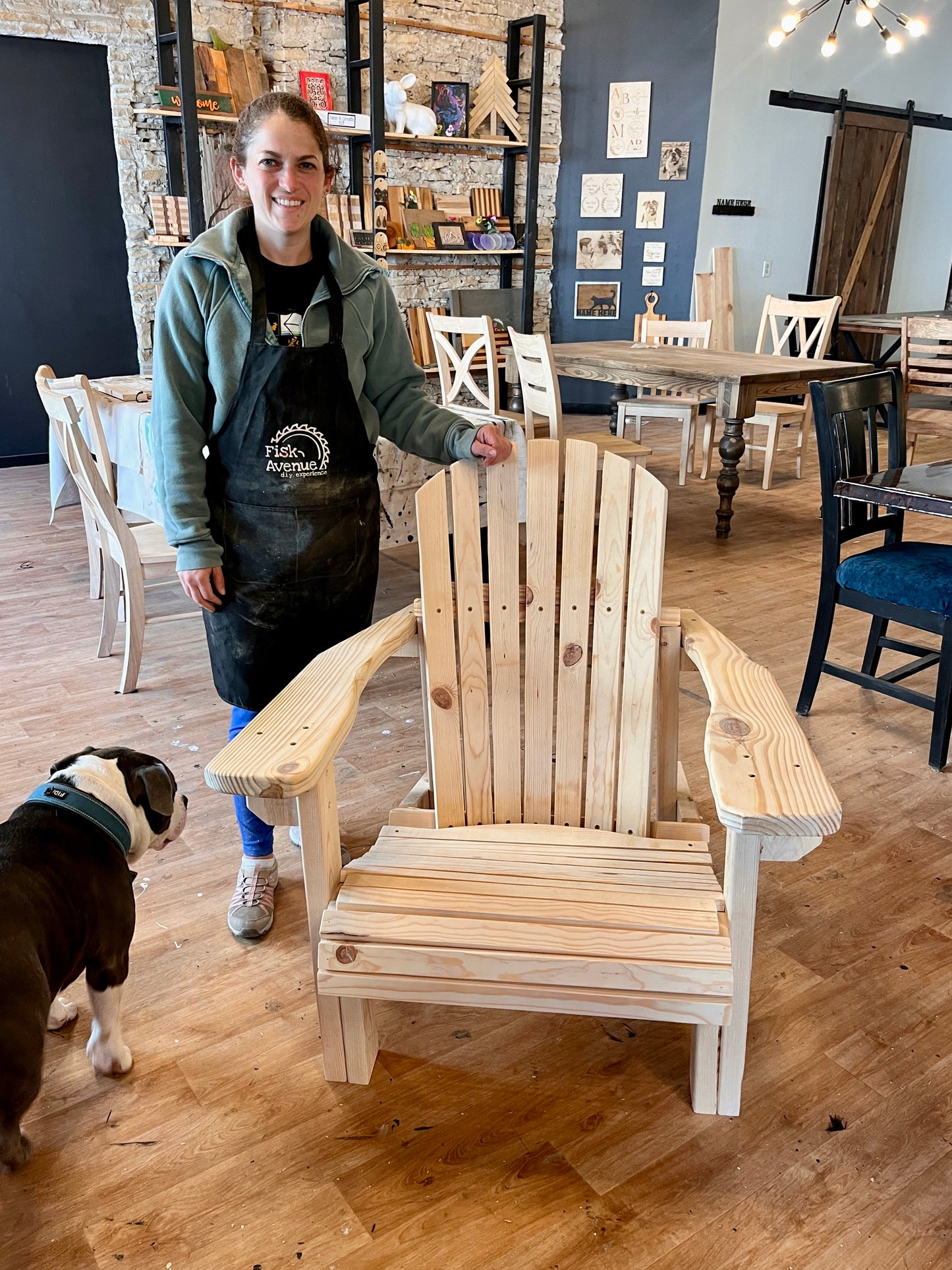 Woman in apron standing with a wooden Adirondack chair in a workshop with a dog.