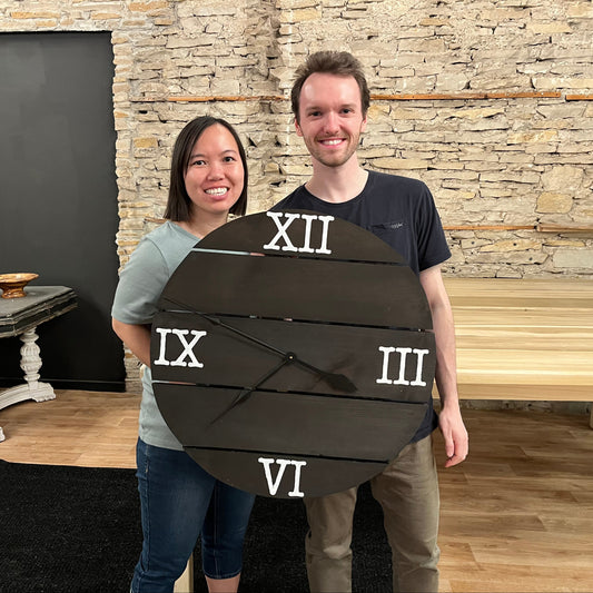 Two people holding a large decorative Roman numeral clock against a stone wall.