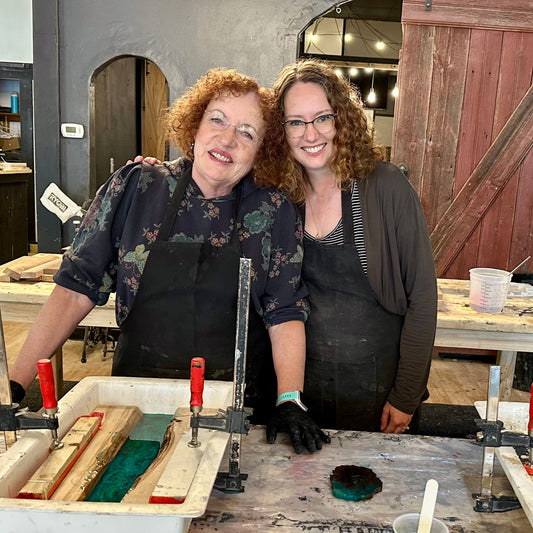 Two women in a workshop setting with tools and materials for epoxy river serving trays on a table.