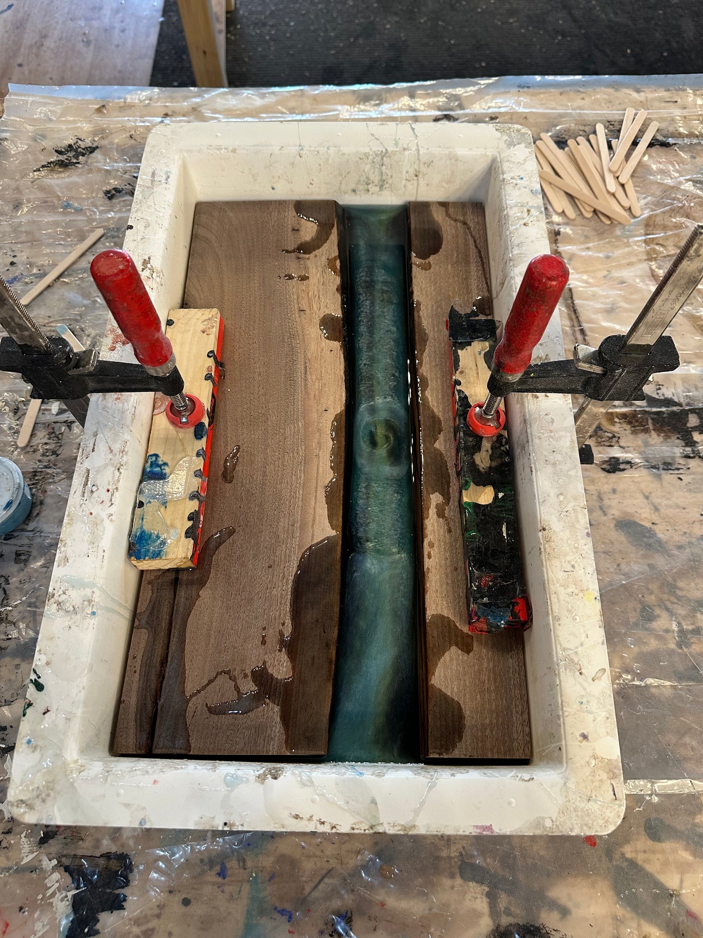 Plastic mold with clamps holding an epoxy river table project with two pieces of live edge wood and an epoxy center on a workshop background
