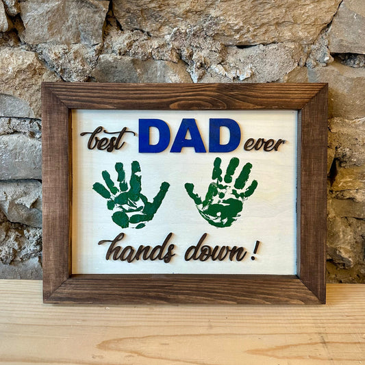 Framed sign with handprints and text 'best DAD ever hands down!' against a stone wall background