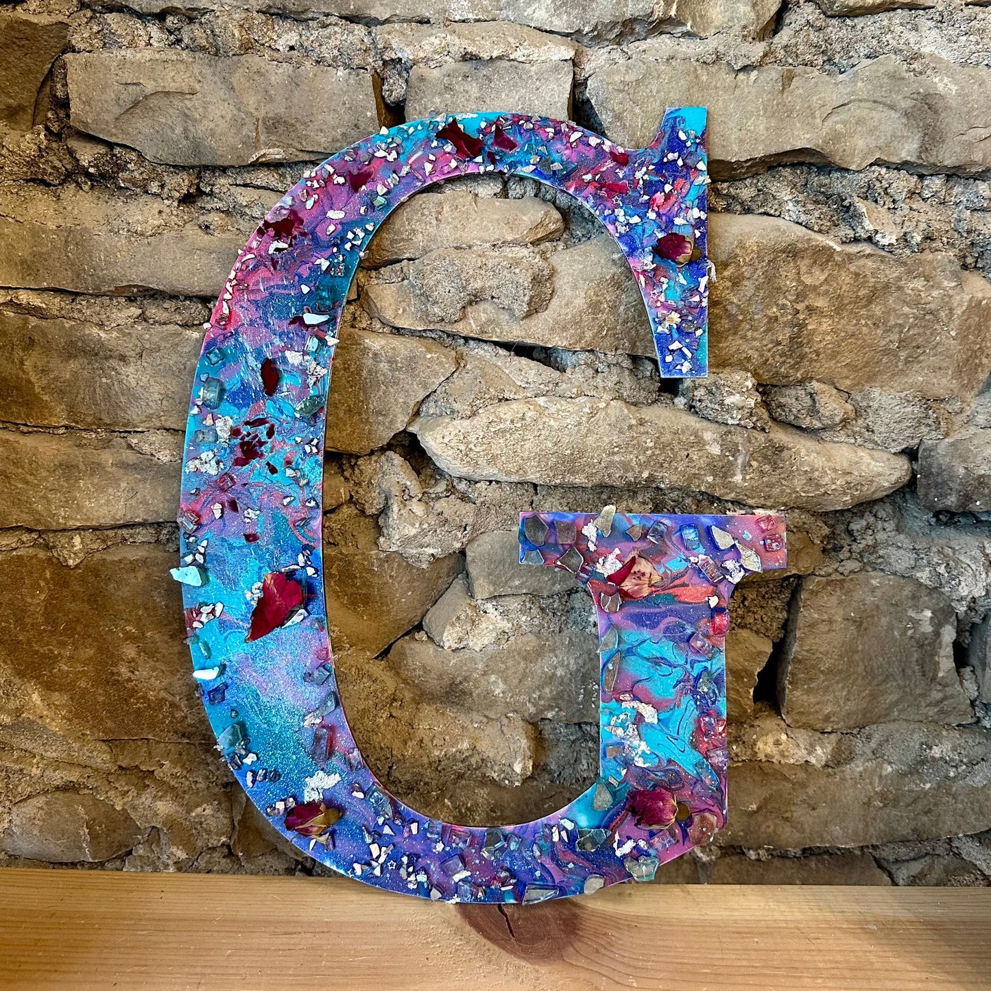 Decorative letter 'G' with a multicolored acrylic pour painted pattern, crystals, and flower petals against a stone wall background