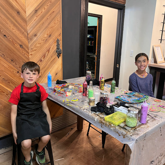 Two children sitting at a table with acrylic pour painted letters in a studio room.