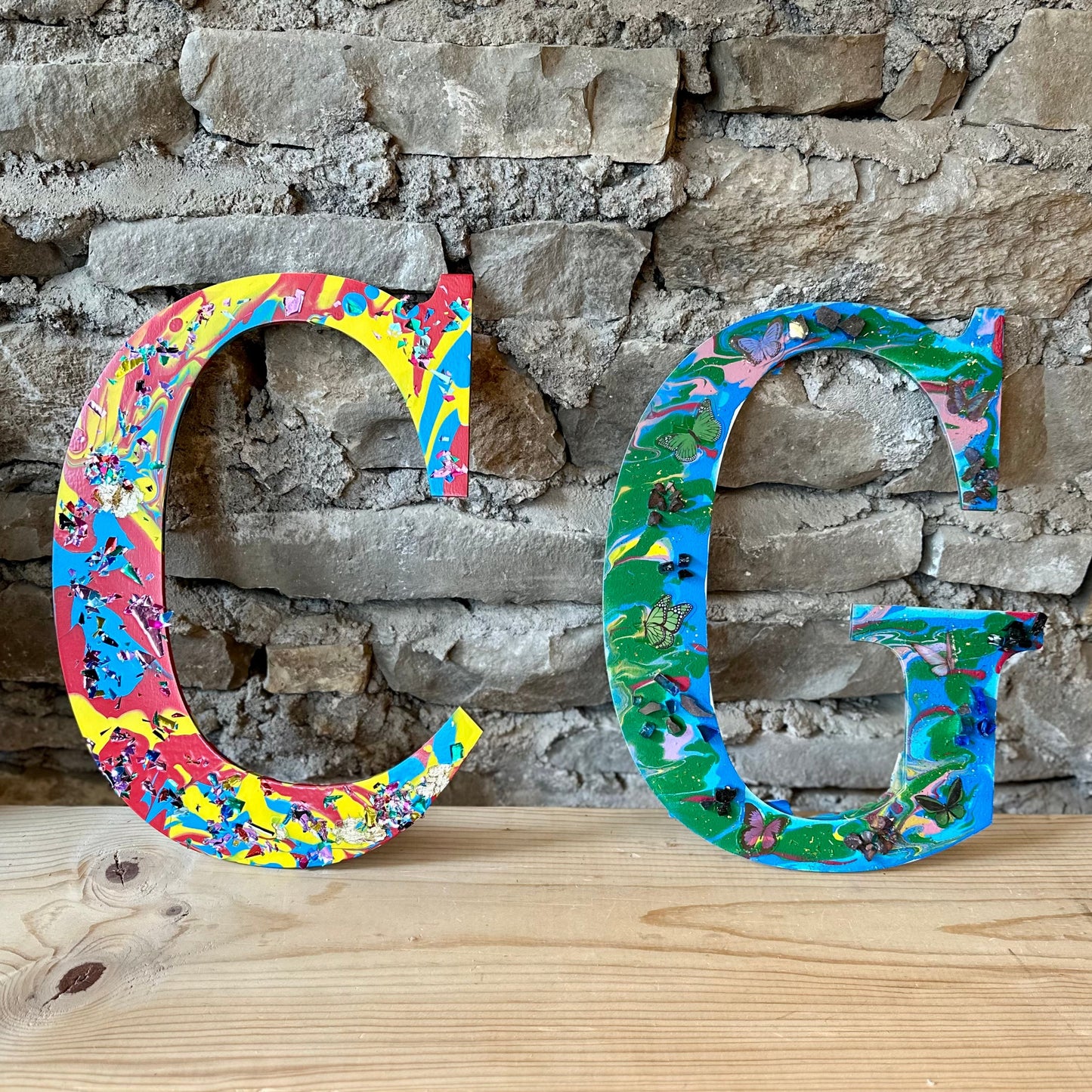 Colorful acrylic pour painted letters 'C' and 'G' with crystals and butterflies on a wooden shelf against a stone wall.