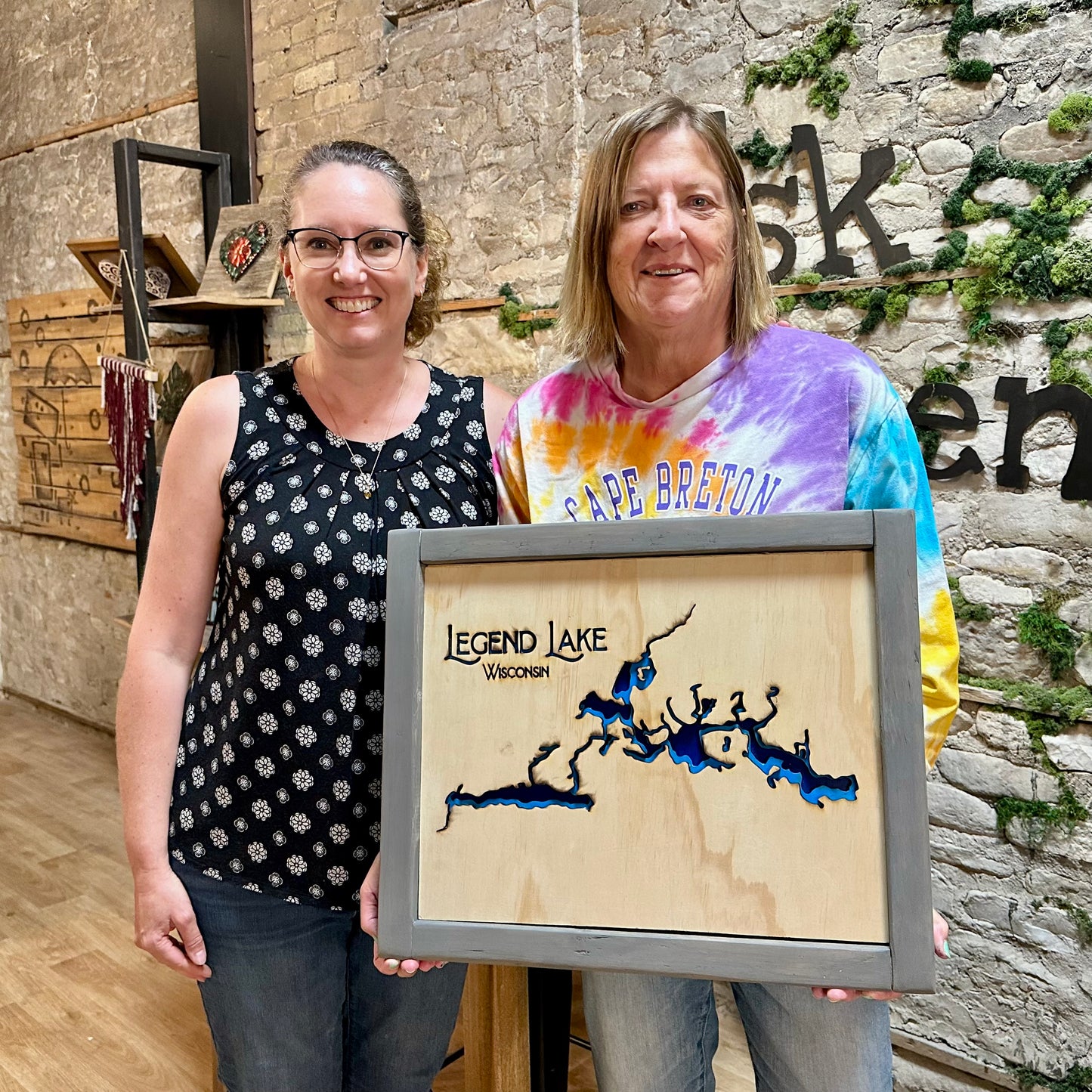 Two women holding a framed wooden depth chart map craft of Legend Lake, Wisconsin against a stone wall background.