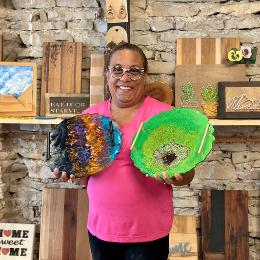 Woman holding two colorful epoxy geode serving trays against a stone wall with various wooden crafts.