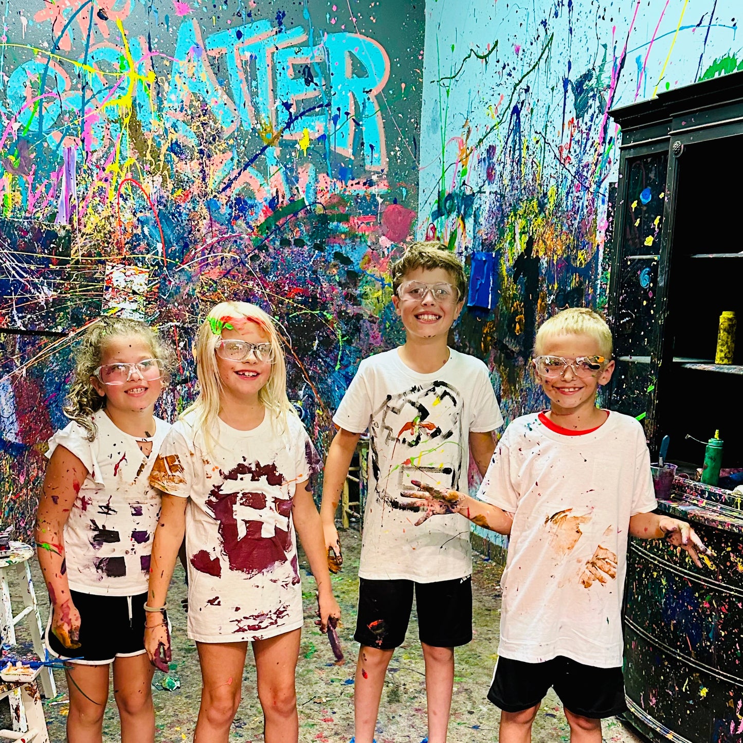 Four children in a splatter room with colorful graffiti on the walls, wearing paint-splattered shirts.