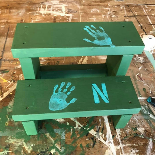A green step stool with a child's handprint and the letter 'N' painted on it, indicating it is a custom-painted product from a workshop.