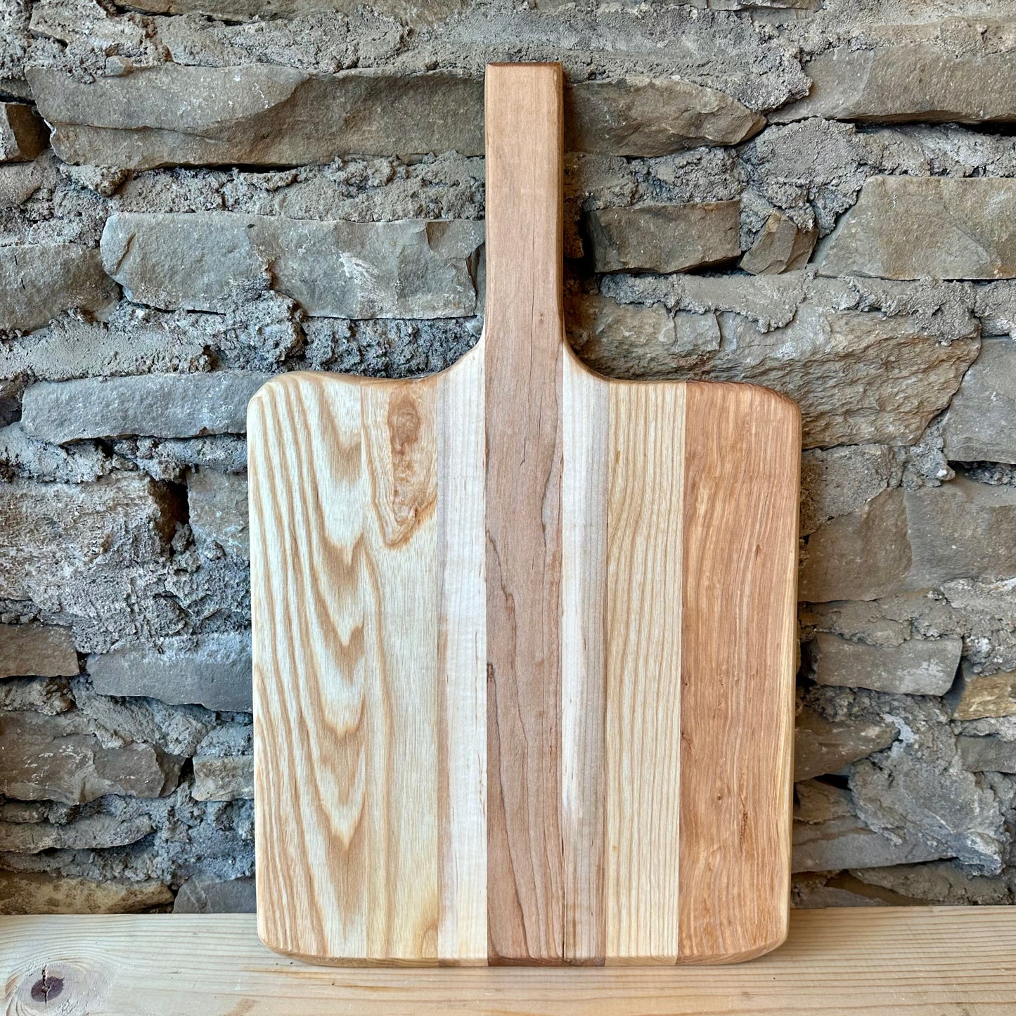 Wooden pizza peel board with a handle on a wooden surface with a stone wall background