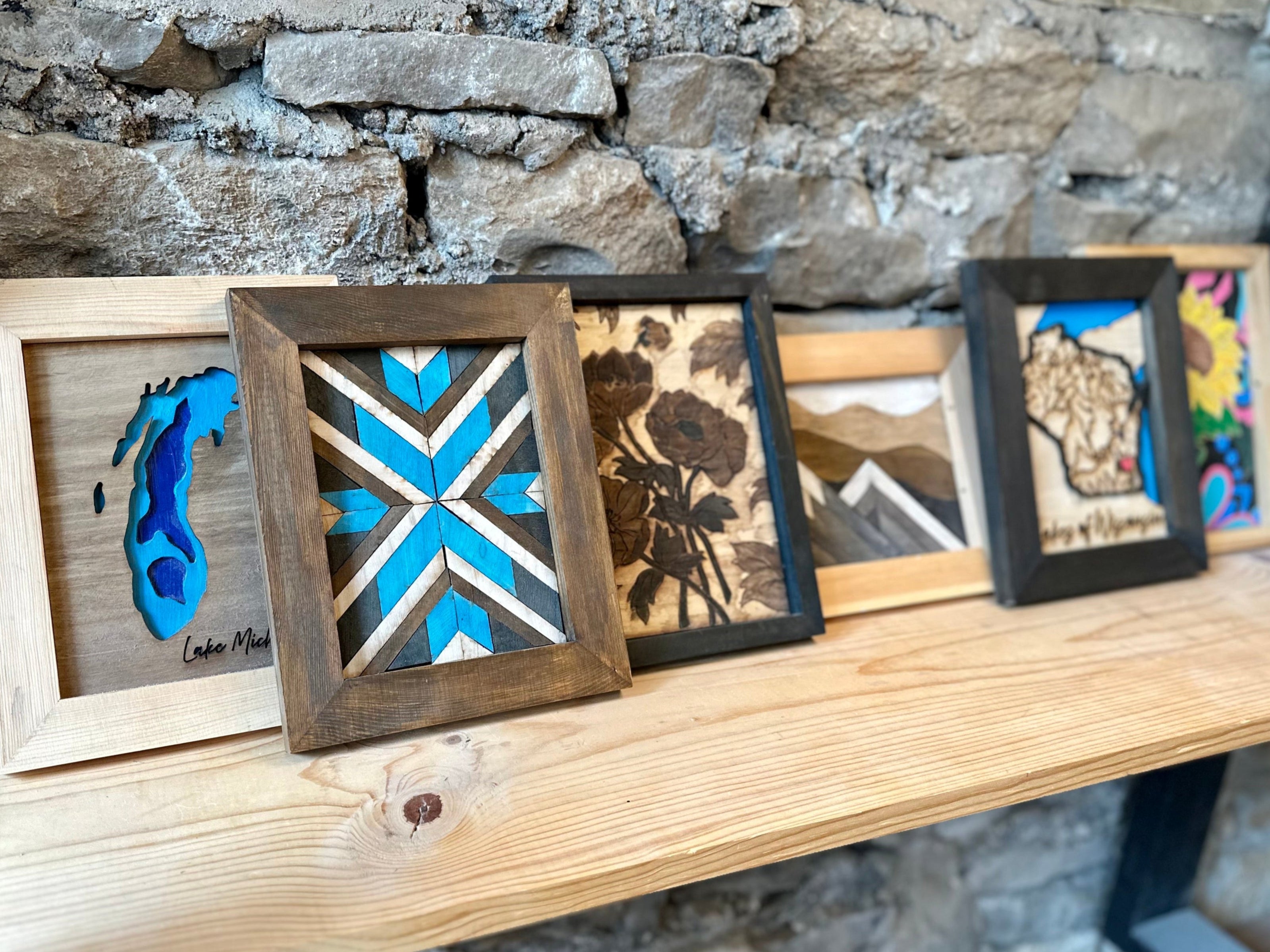 Collection of framed wood art kits on a wooden shelf against a stone wall.